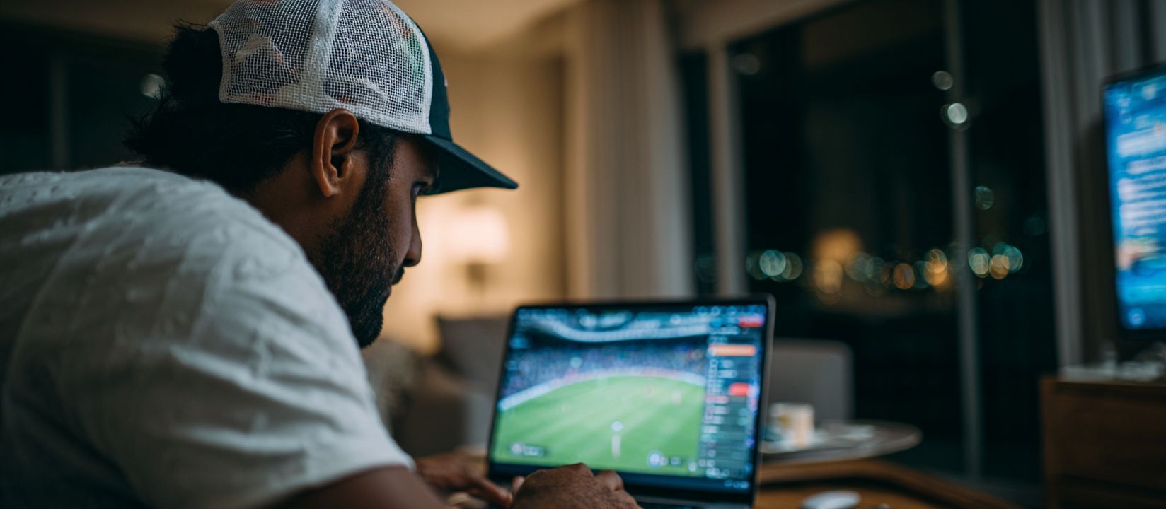 Man wearing a cap placing live football bets on OKLA sports betting platform from his laptop at home